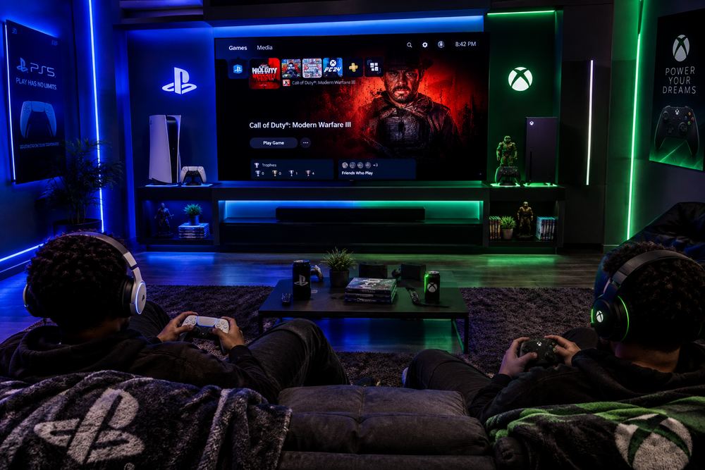 Two players on a sofa with consoles in front of a blue and green lit wall